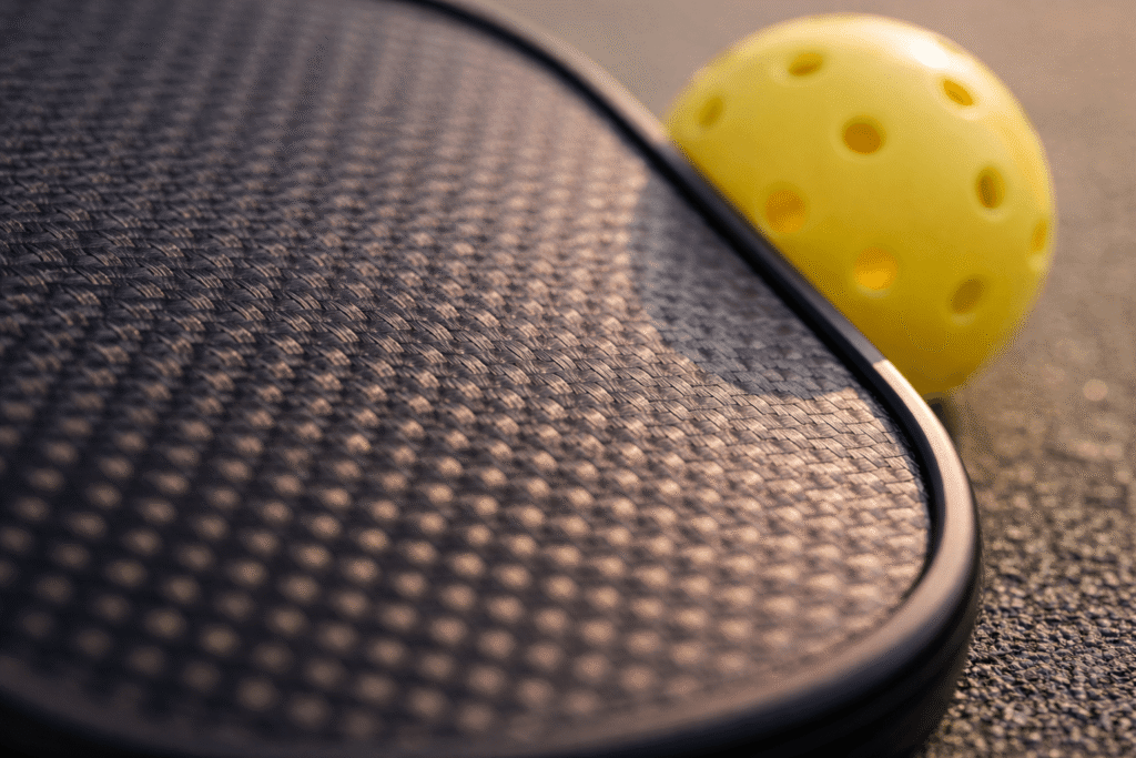 Macro close-up of a raw carbon fiber pickleball paddle face with visible gritty texture and a pickleball resting against the surface